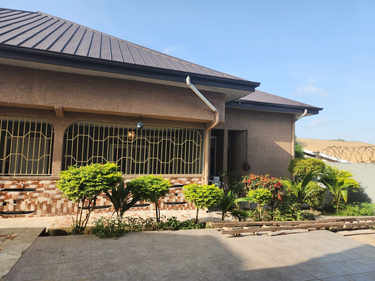 The exterior of a modern home is showcased, featuring a sloped roof and large, arched windows. A manicured garden with vibrant plants frames the entrance, while a pathway extends toward the house, creating an inviting approach.