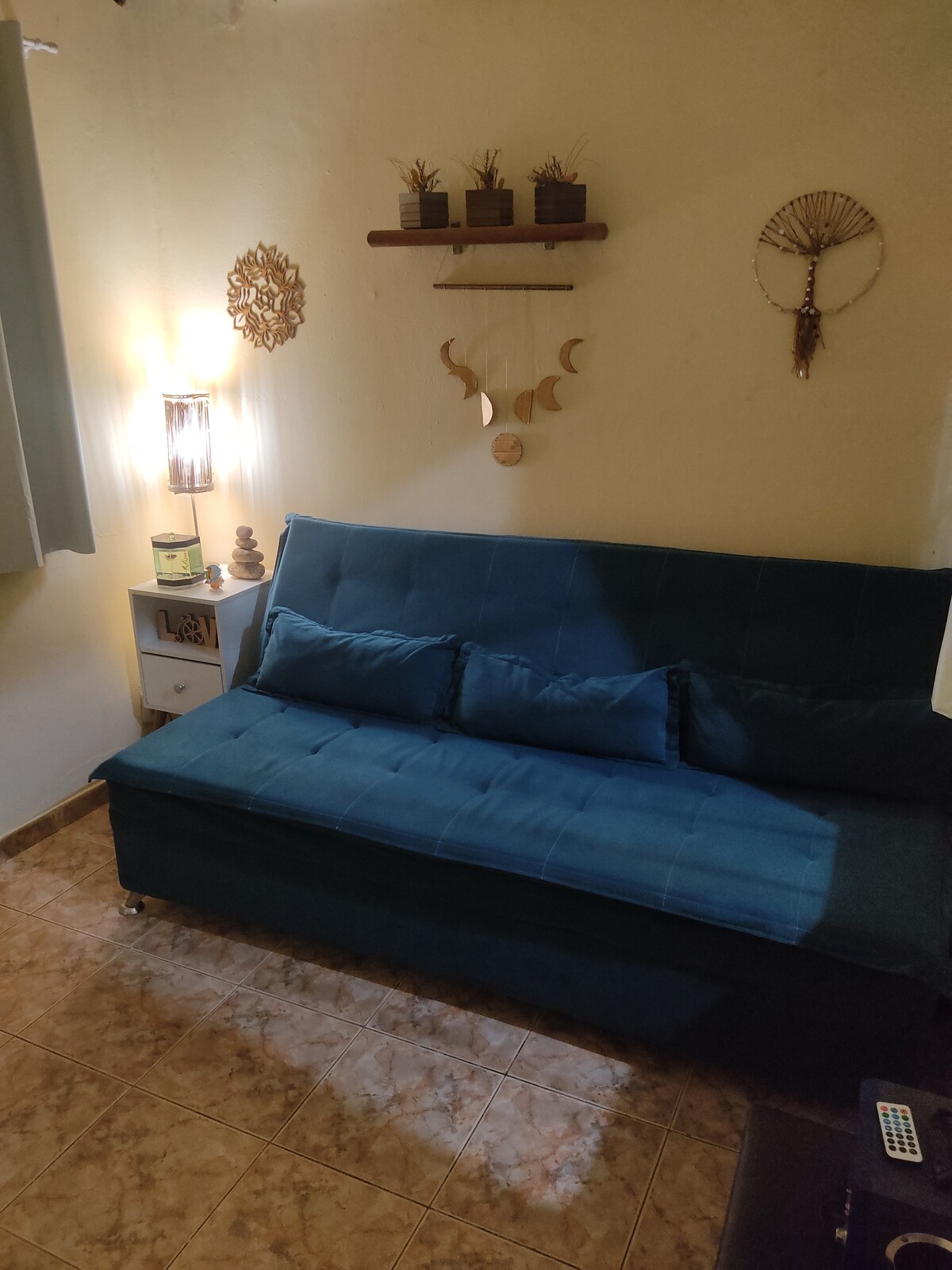 A cozy seating area is presented with a deep blue sofa bed adorned with plush cushions. Wall decor includes potted plants on a shelf, decorative dreamcatchers, and an artful light fixture, contributing to a calming ambiance. Soft gray curtains frame a window, allowing natural light to enter.