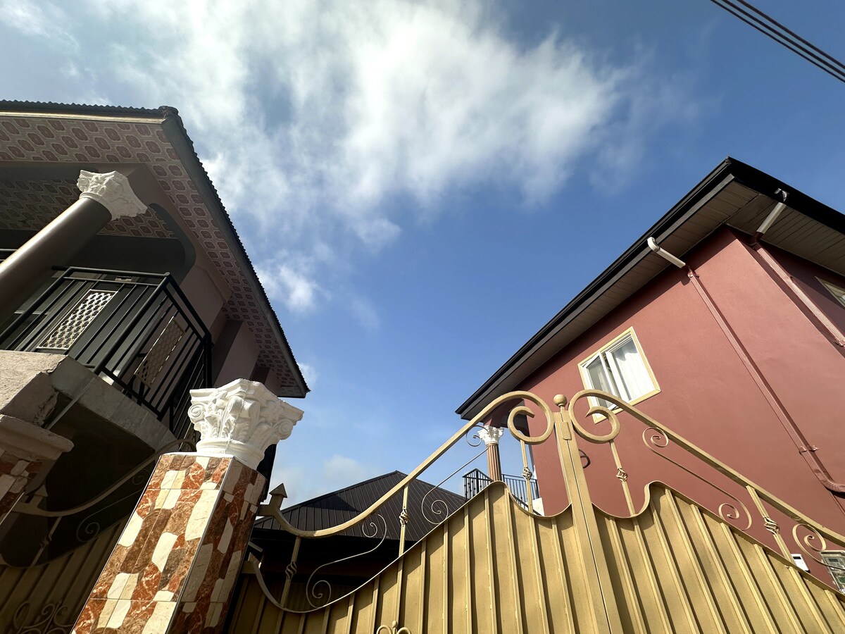 A view shows the entrance gate of the property, framed by ornate pillars. The sky is bright blue with scattered clouds. Two buildings can be seen on either side, one displaying intricate architectural details and the other featuring a simple facade.
