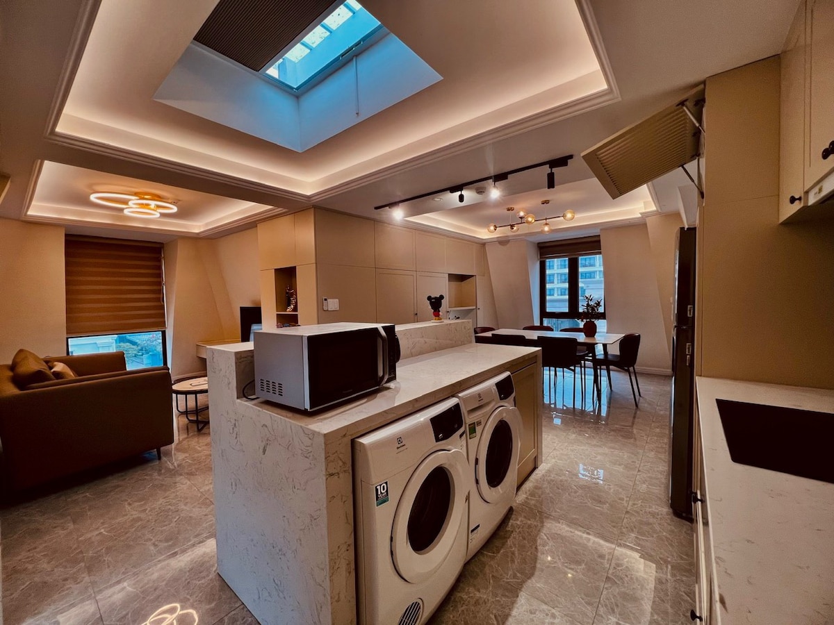 A modern living space features a sleek kitchen island with a microwave and a washer-dryer unit. Natural light filters through a skylight and large windows, illuminating the open layout that includes a comfortable seating area and a dining space.
