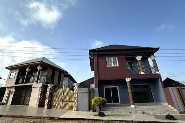 An elegant two-story villa is presented, featuring a welcoming facade with decorative columns and large windows. A manicured shrub is positioned near the entrance. The building is flanked by another structure, showcasing varied architectural styles. A clear blue sky complements the scene.