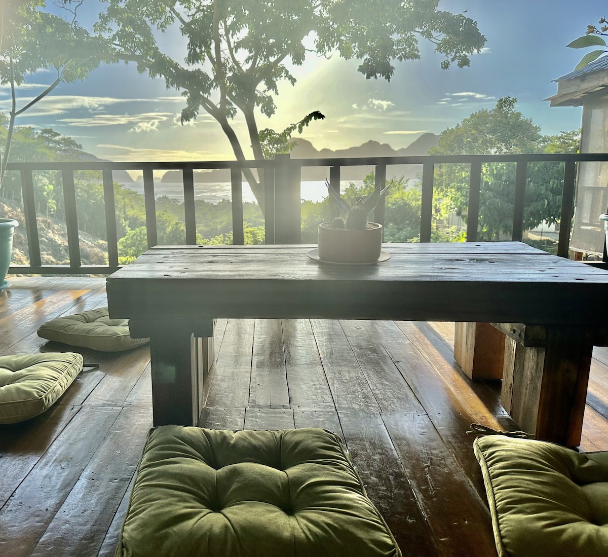 An outdoor dining area is shown with a wooden table in the foreground and cushioned seating arranged around it. Lush greenery and a view of the distant islands are visible through the slatted railing, with soft morning light filtering through the trees.