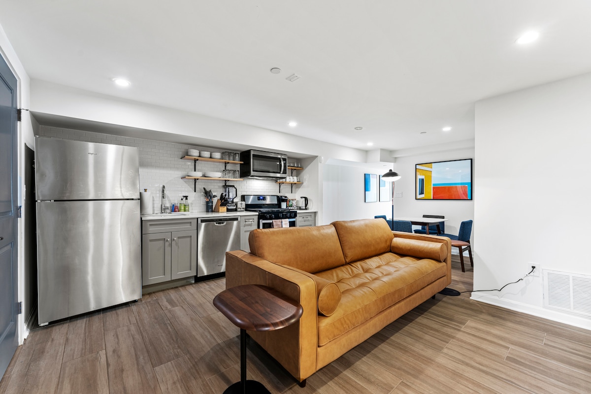 A cozy living area features a brown leather sofa facing a modern kitchen area, equipped with stainless steel appliances and open shelving. To the side, a dining table with blue chairs adds a pop of color, complemented by framed artwork on the walls.