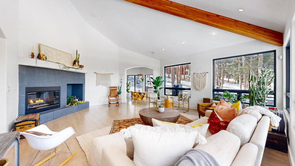 An open living area features vaulted ceilings and a modern gas fireplace. Large windows provide ample natural light and views of the surrounding nature. Plush seating is arranged around a circular coffee table, with greenery creating a fresh atmosphere.