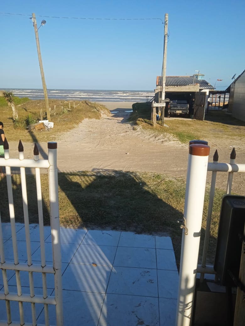 A view from the entrance gate opens to a sandy path leading toward the beach. The path is bordered by grass on either side, and utility poles stand nearby. A vehicle can be seen parked under a structure to the right, with the ocean visible in the background.