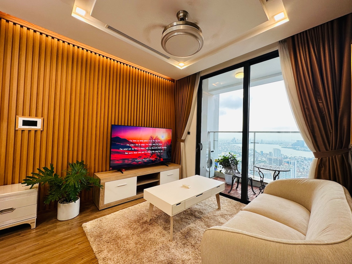 A bright living room features a large smart TV against a wooden accent wall. A light-colored sofa sits adjacent to a coffee table on a plush area rug. Floor-to-ceiling windows offer a view of the cityscape, adorned with warm curtains framing the space.
