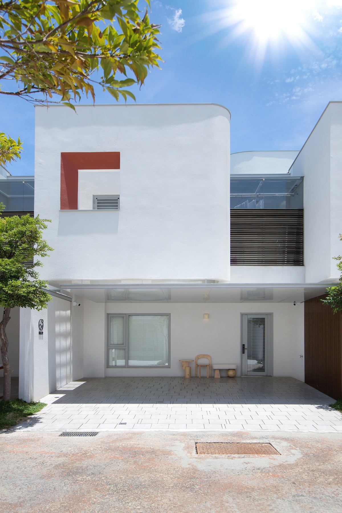 The exterior of a modern building is presented, featuring a minimalist design with smooth white walls and large windows. A small seating area with two wooden chairs is positioned in front, framed by greenery. Bright sunlight casts a warm glow over the entrance.