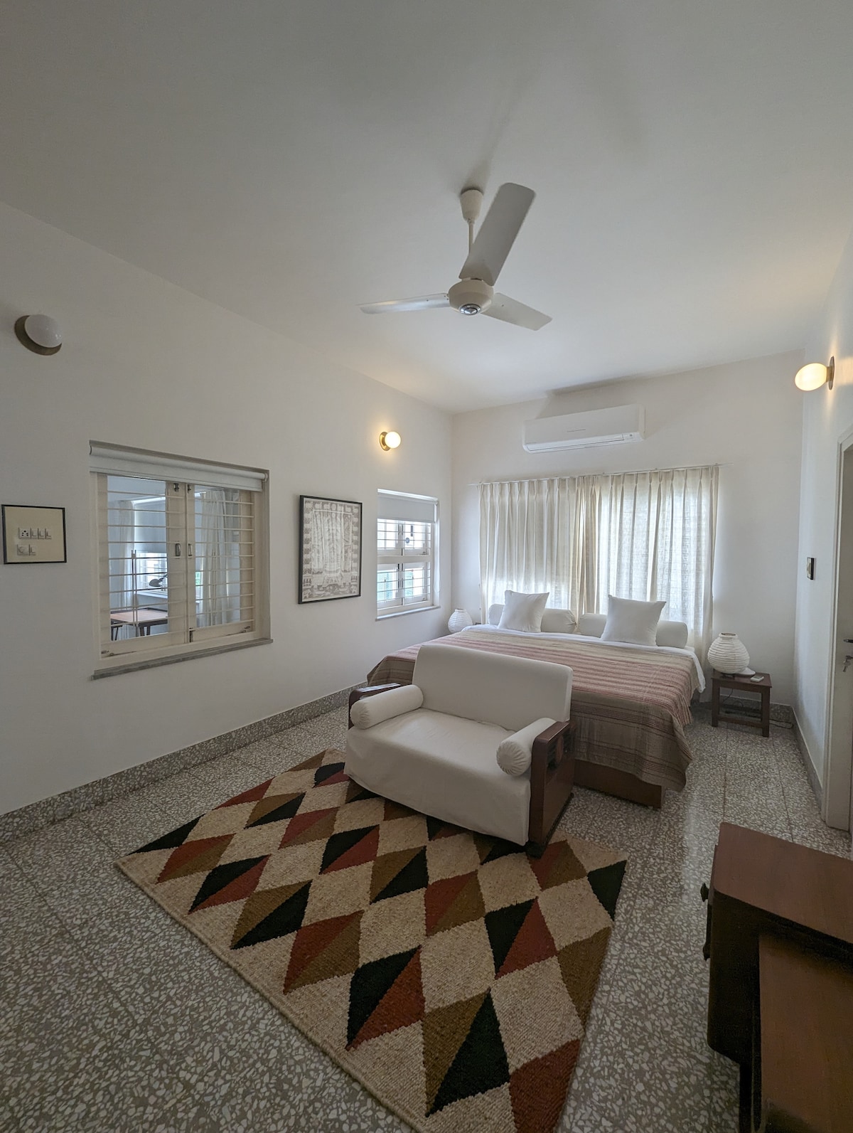 A spacious bedroom features a large bed with light-colored linens and two bedside lamps. A comfortable white armchair is placed near a geometric patterned rug. Natural light filters through sheer curtains, illuminating the room's soft tones and creating a cozy ambiance.