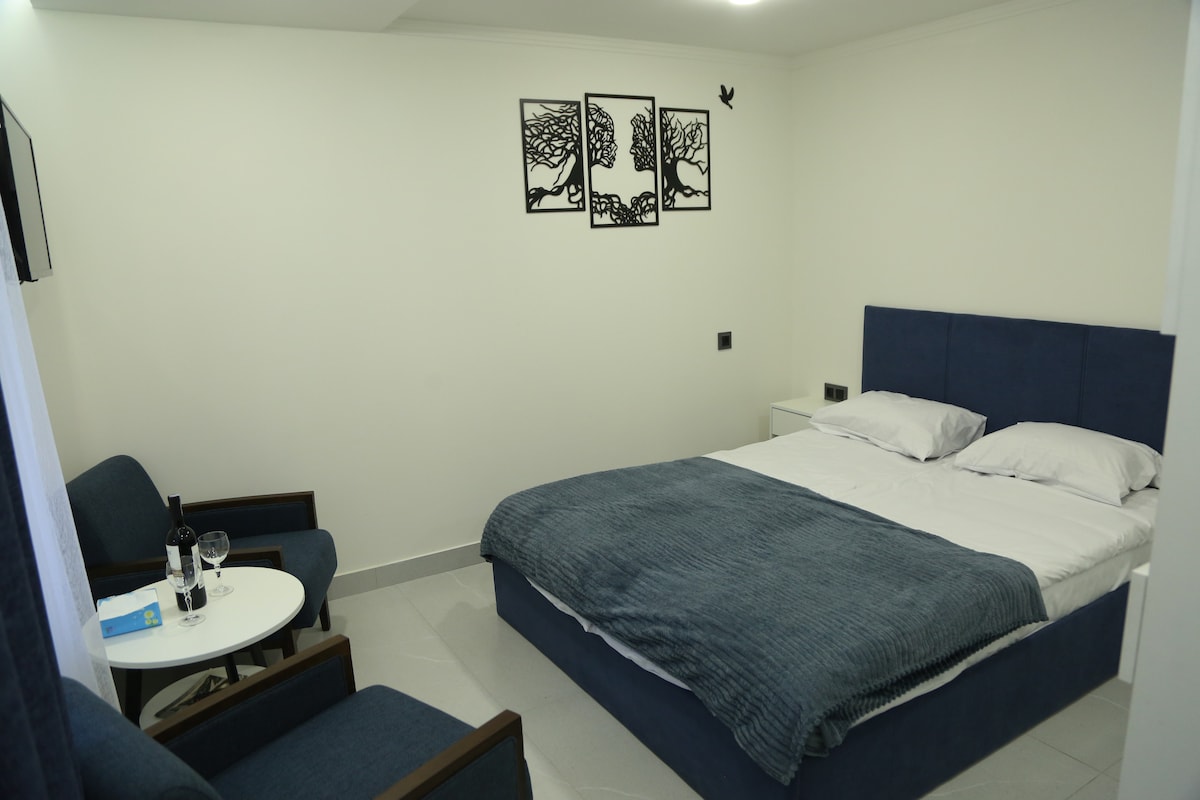 A well-arranged bedroom features a spacious bed with a soft blue blanket. Two dark armchairs and a small white table can be seen in one corner. Wall art depicting trees adds a touch of interest above the bed, while the space is illuminated by soft lighting.