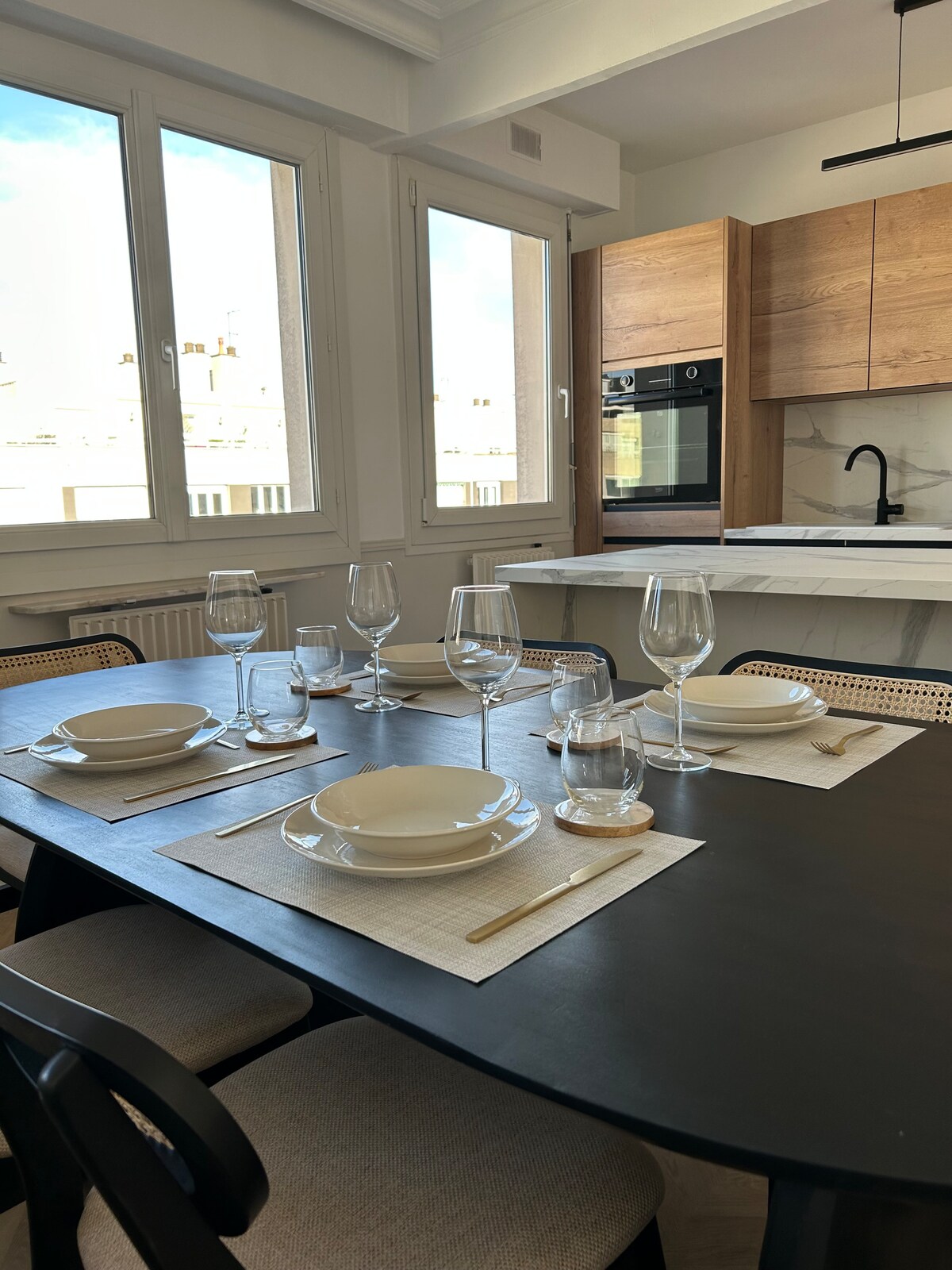 A modern dining area features a dark table set for six, complemented by white plates, crystal glassware, and simple placemats. Natural light floods the space through large windows, highlighting the sleek cabinetry and contemporary appliances of the kitchen in the background.