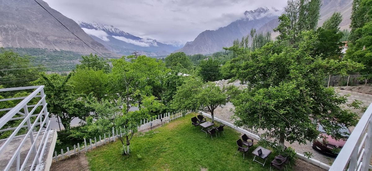 A spacious outdoor area is surrounded by lush greenery, with several seating arrangements set under the shade of trees. The landscape features distant mountains shrouded in clouds, providing a serene backdrop to the garden space.