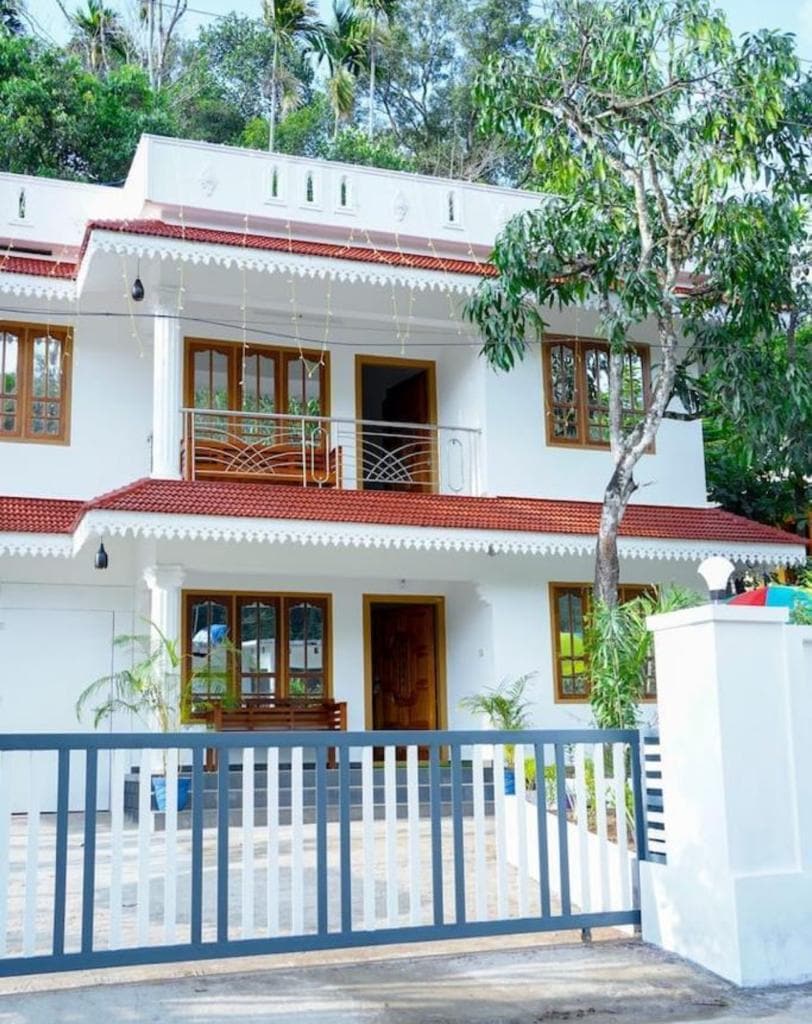 A charming two-story villa is framed by a landscaped garden and surrounded by tropical trees. The light-colored exterior features multiple large windows with wooden accents and orange tiles on the roof. A gated entrance leads to the property, offering a sense of privacy.