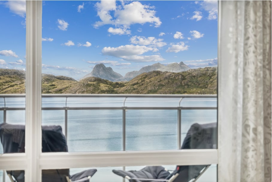 A serene view of the ocean and rolling mountains is framed by large glass doors. Two chairs, positioned on the balcony, invite relaxation while curtains softly diffuse the light. The landscape features clear blue skies and calm waters, enhancing the peaceful ambiance.