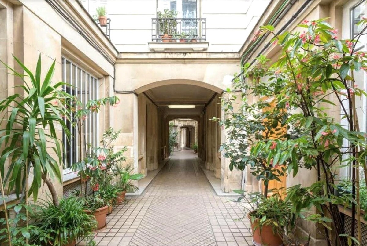 An inviting courtyard is bordered by lush greenery and potted plants, leading to archways that enhance the sense of space. Natural light filters through the openings, creating a serene atmosphere in the heart of the residence.