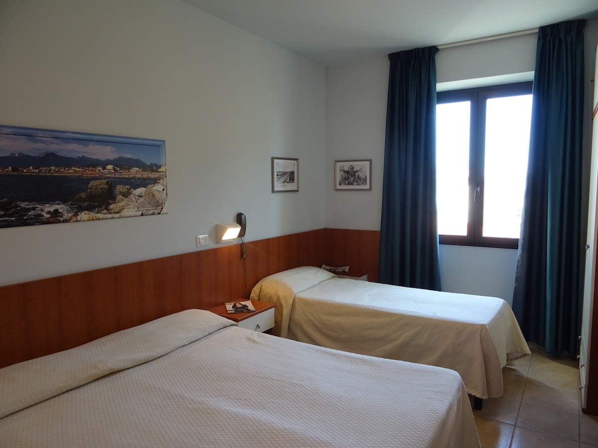 A well-lit room features two spacious beds covered with light bedding. A window is positioned on one wall, allowing natural light to enter. Dark curtains frame the window, while framed pictures adorn the walls, providing a touch of decor.