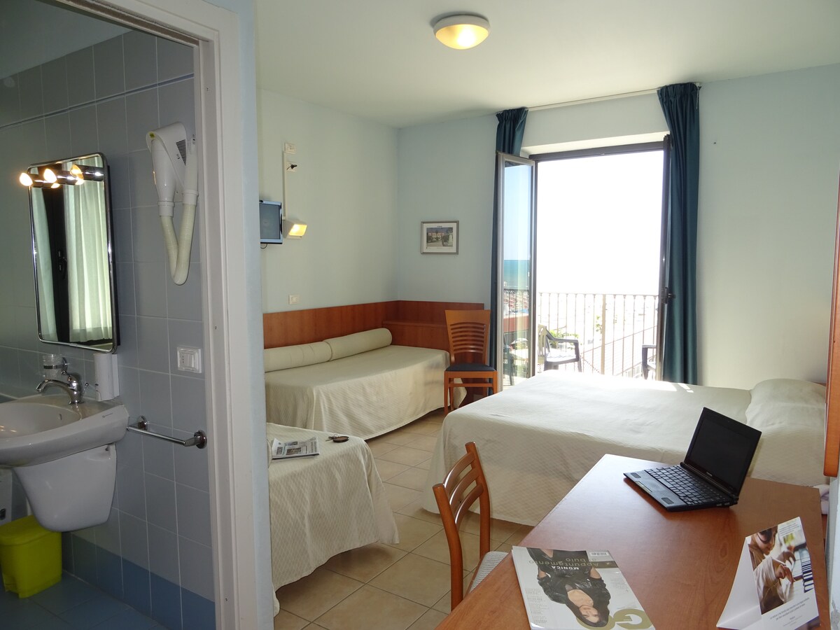 A well-lit room features a spacious bed and a separate seating area with two cushioned couches. A desk with a laptop is positioned next to a large window that opens to a balcony, offering views of the sea. A bathroom is visible with simple fixtures.