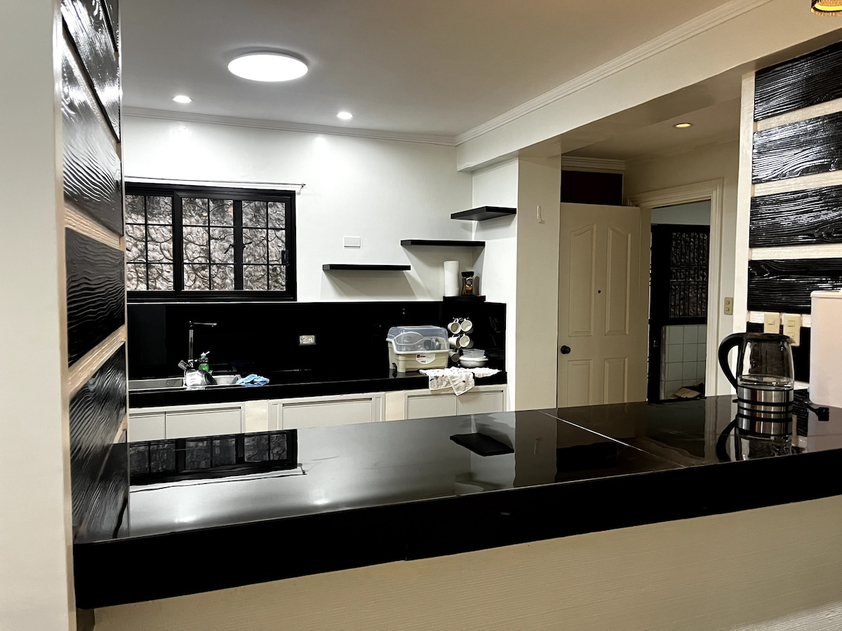 A modern kitchen showcases sleek black countertops and white cabinetry. Open shelving lines the wall, providing space for kitchen essentials. Natural light pours in from a multi-paned window, enhancing the functionality of the space. A kettle is positioned on the counter for convenience.