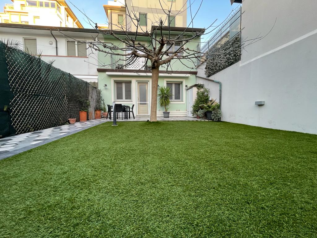 A spacious private garden features vibrant green artificial grass, bordered by a low fence. A tree stands in the center, and a set of outdoor furniture, including a table and chairs, is positioned nearby. The exterior of the building is visible in the background.
