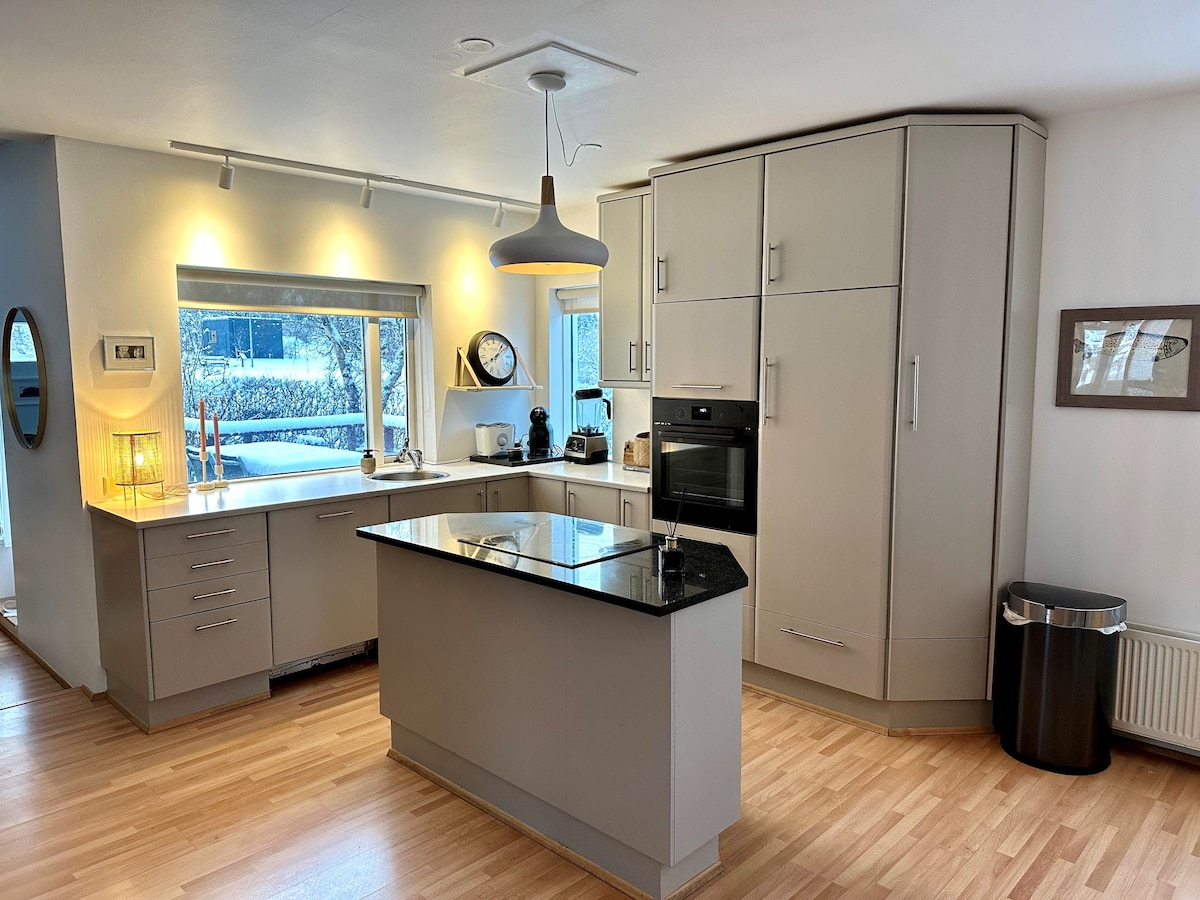 The modern kitchen features sleek cabinetry and an island with a contrasting dark countertop. Aromatic appliances, including an oven, are integrated into the design. A window allows natural light to brighten the space, while wooden flooring adds warmth to the environment.
