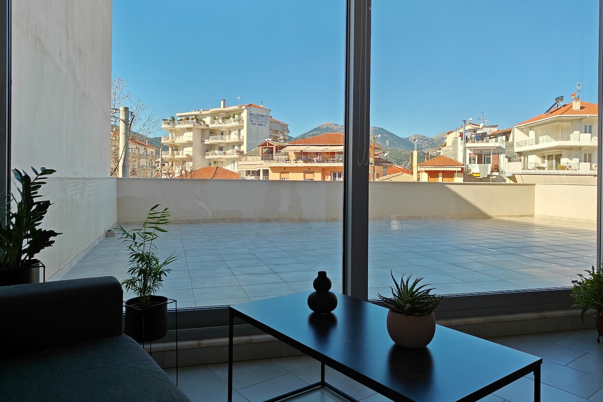 A spacious balcony is visible through large glass doors, featuring a table and plants. The view includes surrounding buildings and distant mountains under a clear blue sky, creating a bright and open atmosphere.