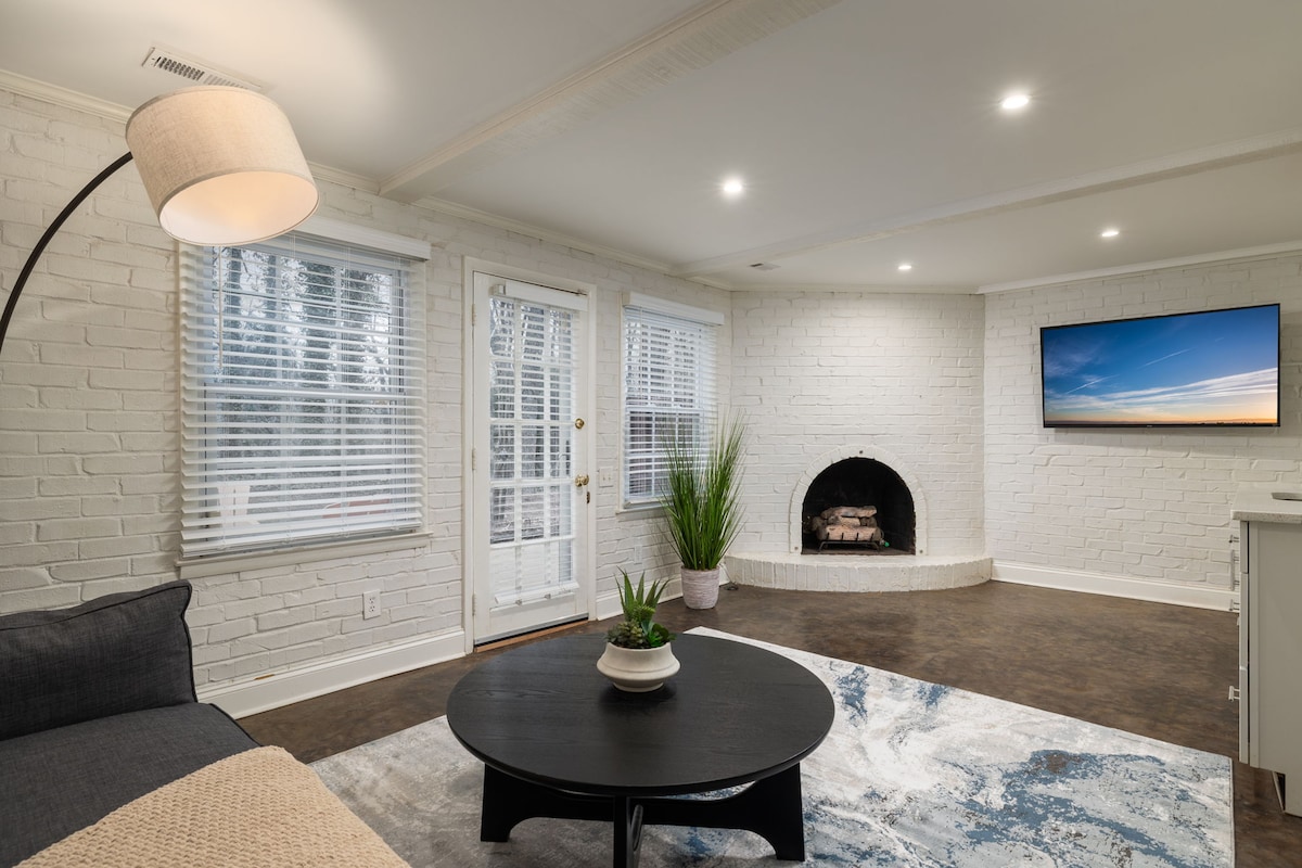 A spacious living area features a modern grey couch beside a round black coffee table. A decorative plant is centered on the table. A traditional fireplace is framed in the background, with a flat-screen TV mounted above it. Natural light filters through the white, vertical blinds.
