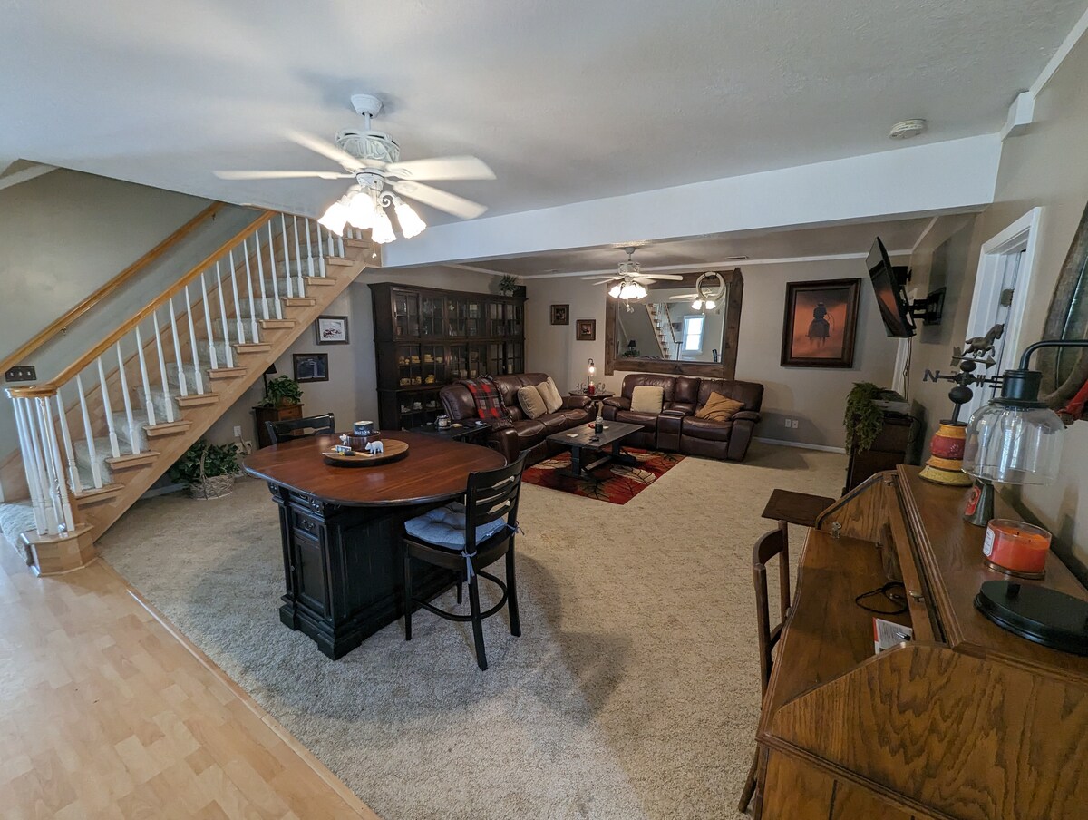 A spacious living area is highlighted by a centrally placed wooden dining table surrounded by chairs. Comfortable seating is arranged around a coffee table on a soft carpet. A staircase is visible, leading to the upper level, while a ceiling fan provides a sense of airflow.