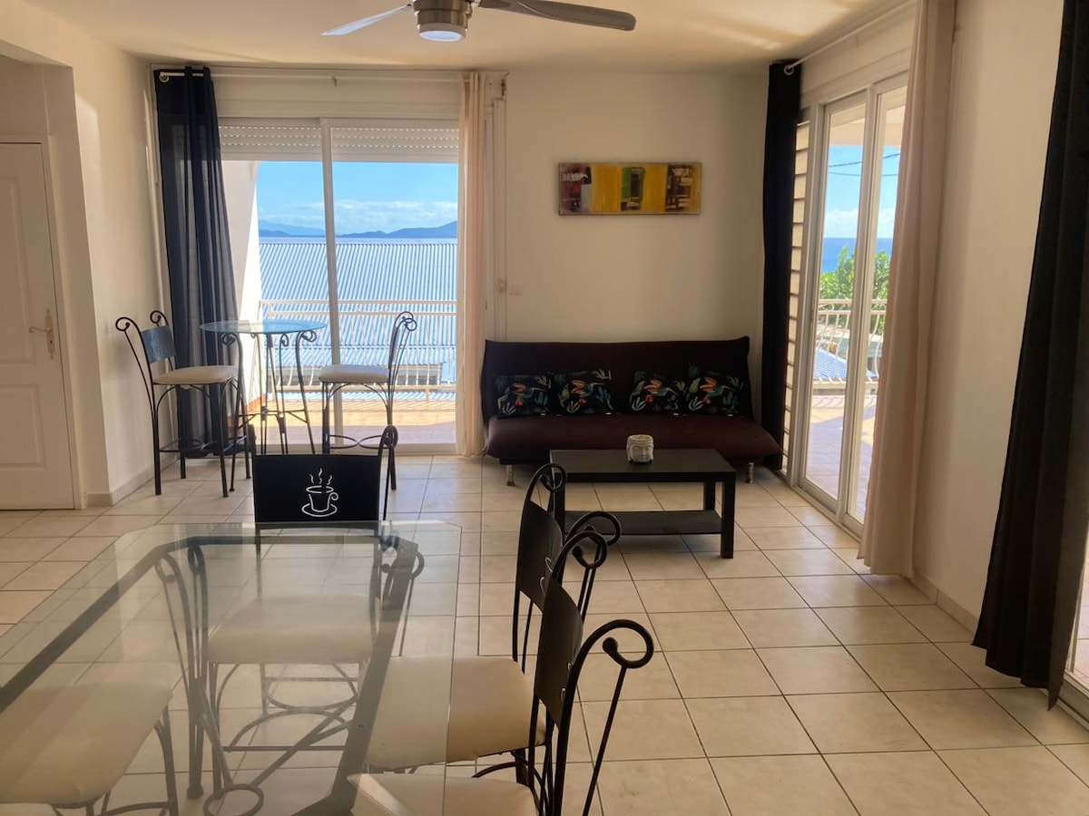 A bright living area features a glass dining table surrounded by six chairs. Large sliding glass doors offer views of the outdoor space and ocean. A dark sofa with colorful cushions complements the neutral decor, while a ceiling fan provides airflow.