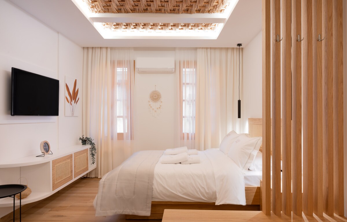 A modern bedroom features a spacious bed dressed in crisp white linens. Natural light filters through sheer curtains, illuminating wooden accents and wall art. A flat-screen television is mounted on the wall, and wooden partitions add a touch of privacy.