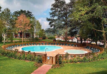 A circular swimming pool surrounded by a wooden fence is set within a lush, green garden. A shallow kiddie pool is visible adjacent to the main pool. Sun loungers are positioned around the pools, and well-maintained trees provide shade in the area.
