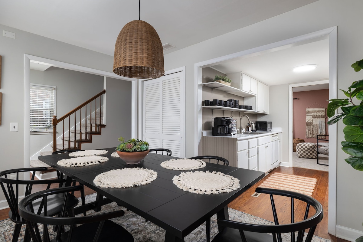 A modern dining area features a large black table surrounded by black chairs, set with natural fiber placemats. A potted plant adds a touch of greenery, while a woven pendant light hangs above. The adjacent open space leads to a well-equipped kitchen.
