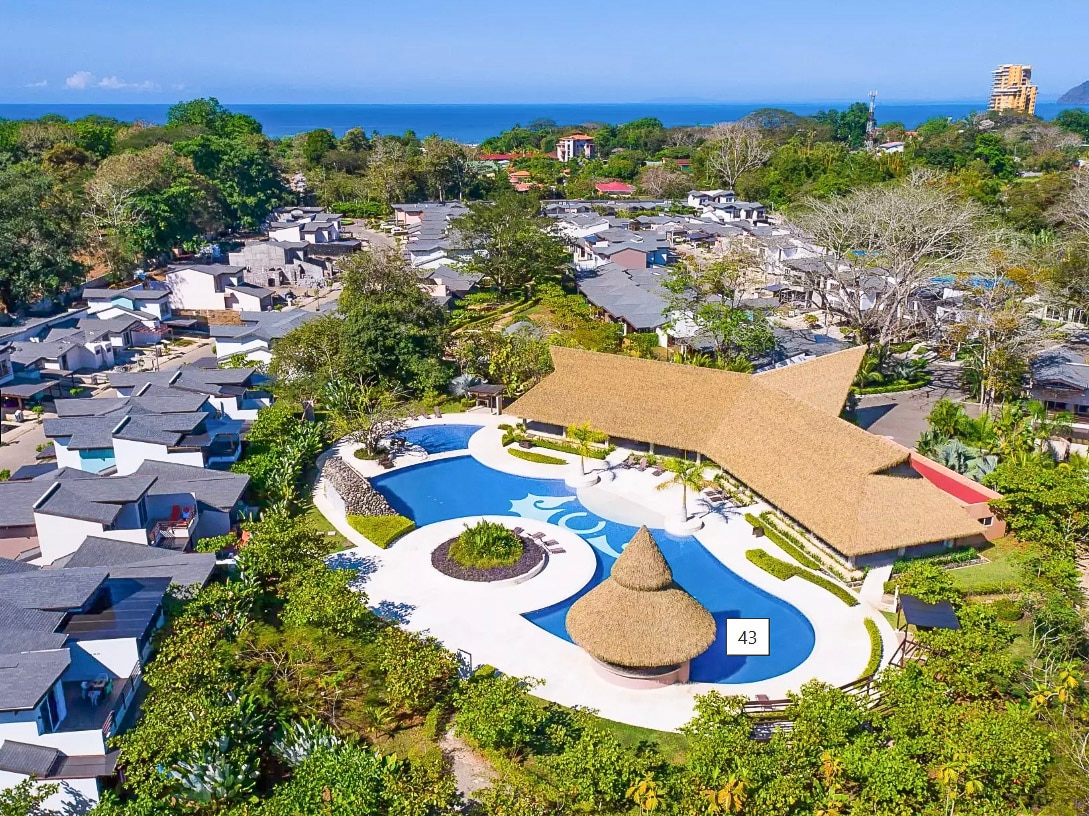 An aerial view captures a resort-style pool surrounded by lush greenery and well-maintained pathways. The open pavilion features a thatched roof, and the ocean can be seen in the distance, providing a serene backdrop to the vibrant resort community.