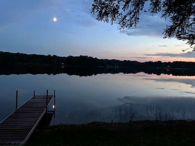 A serene view of Lake Lewis during twilight, featuring a wooden dock extending into the calm waters. The moon reflects softly on the lake's surface, with trees lining the shoreline in the background against a gradient sky transitioning from blue to soft pink.