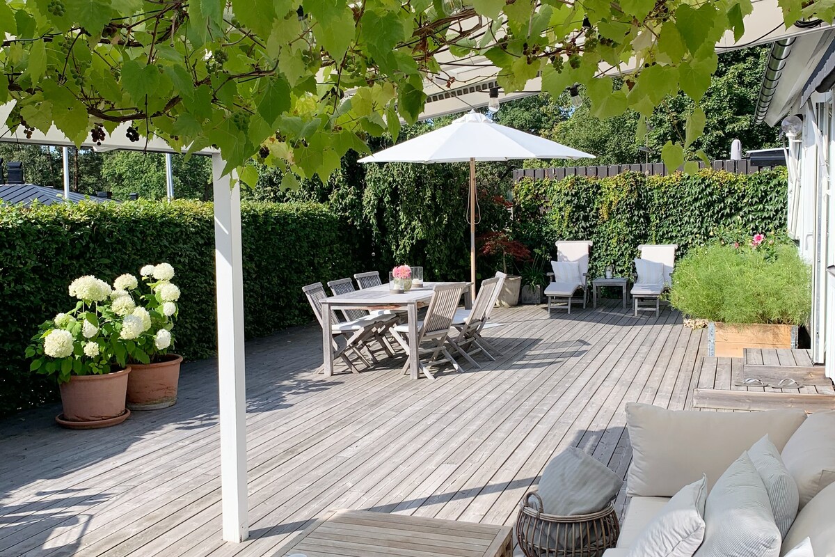 A spacious terrace features a wooden deck surrounded by lush greenery. A large dining table with wooden chairs accommodates outdoor meals. An umbrella provides shade, while potted plants add color and vibrancy to the area. Cozy seating is arranged for relaxation.