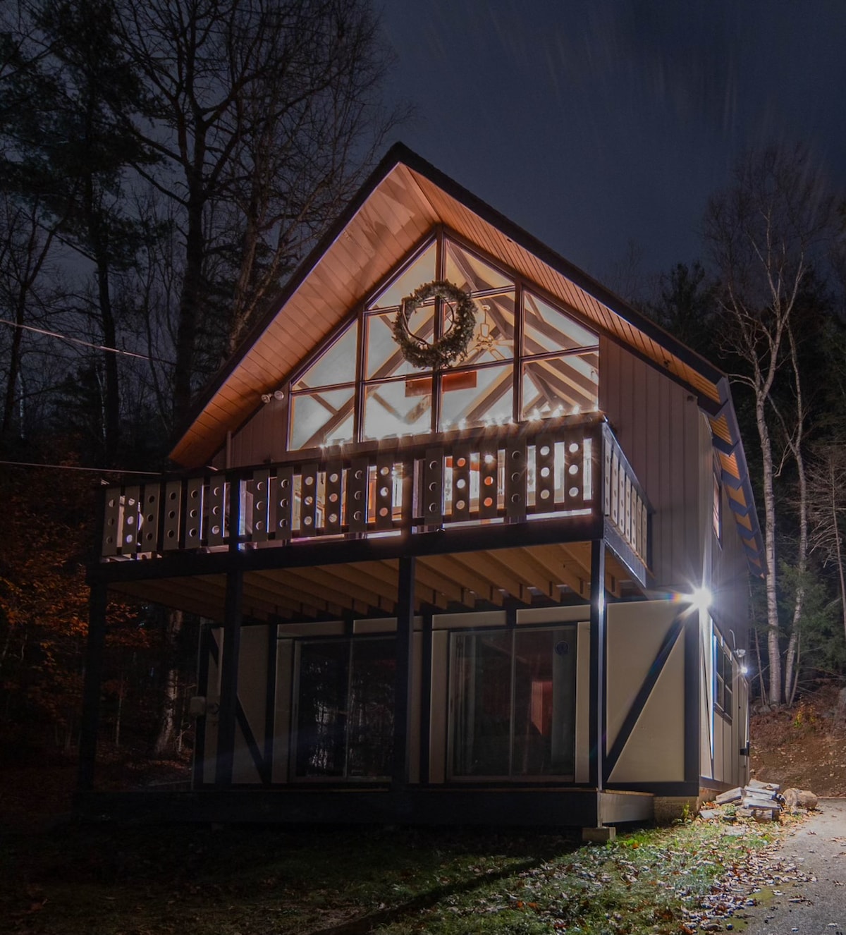 A cozy chalet is presented at night, featuring a striking triangular roof and large glass doors. The balcony is decorated with a festive wreath, and warm lighting highlights the architectural details. Surrounding trees add to the serene atmosphere of the property.