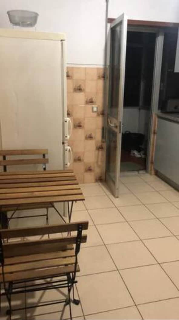 A compact kitchen area features a wooden table with two folding chairs. A refrigerator is positioned against the wall, with light-colored tiled flooring extending throughout. A glass-paned door leads to an adjacent space, adding to the open feel.