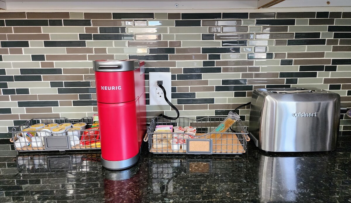A well-appointed kitchen counter features a red Keurig coffee maker alongside a silver toaster. Two wire baskets hold an assortment of coffee pods, utensils, and snacks, all set against a backdrop of stylish multicolored glass tiles.