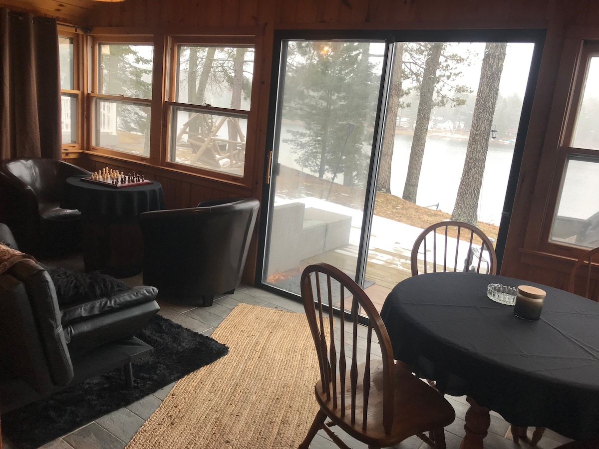 A cozy living area features large windows that provide views of the lake, allowing natural light to fill the space. A black table setting is seen beside a seating arrangement that includes two armchairs and a sofa. A chess set is positioned on a side table.