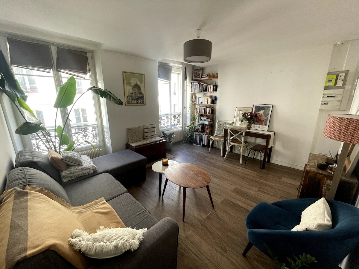 A cozy living area features a gray sofa, a wooden coffee table, and a blue accent chair. Bright light enters through large windows, highlighting bookshelves and decorative elements. Indoor plants add a touch of greenery, creating a welcoming ambiance.