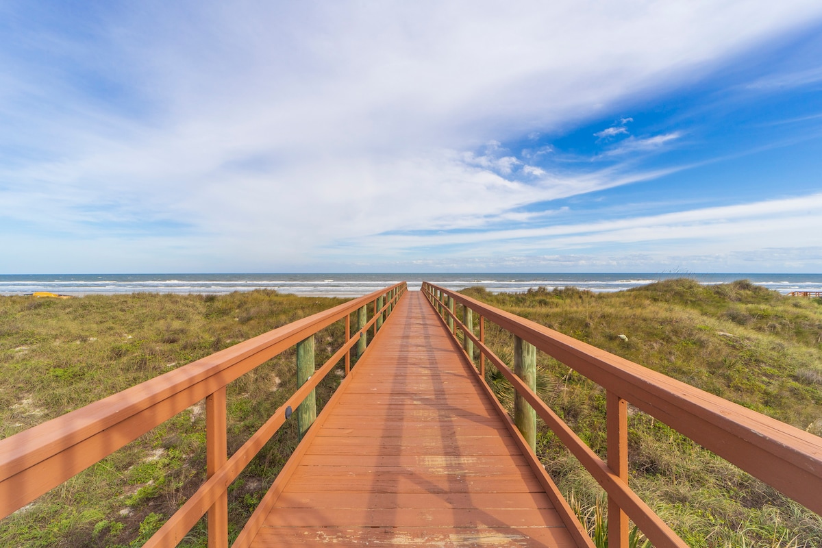 A wooden boardwalk extends toward the ocean, framed by soft green grass on either side. The scene is illuminated by a clear blue sky with scattered clouds, leading to an expansive shoreline where the sea meets the horizon.
