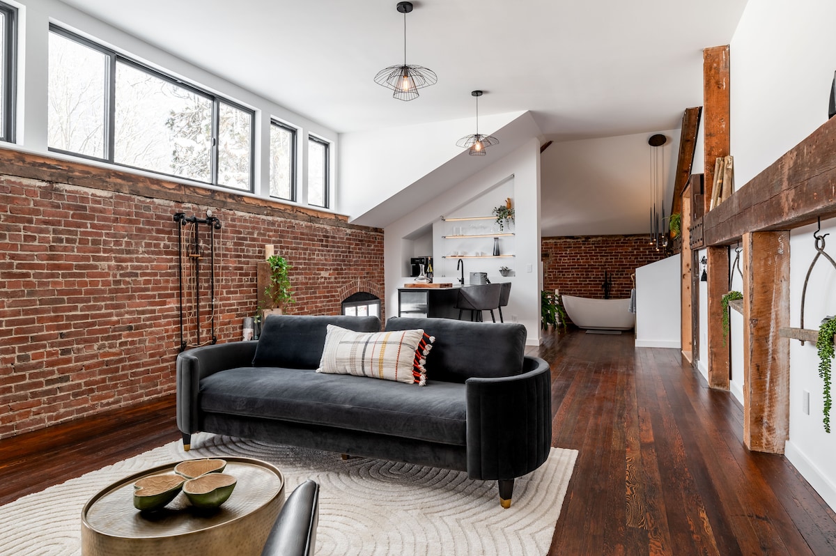 A spacious living area showcases exposed brick walls and large windows that allow natural light to fill the room. A sleek black sofa is positioned on a textured area rug, accompanied by a round coffee table. The stylish kitchenette is visible in the background, with modern appliances and decor.
