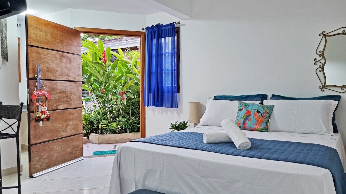The loft features an inviting bedroom with a spacious bed adorned with crisp white linens and decorative pillows. A sliding wooden door leads to a lush outdoor garden, while a vibrant blue curtain allows natural light to illuminate the space. A mirror and small plant accentuate the décor.