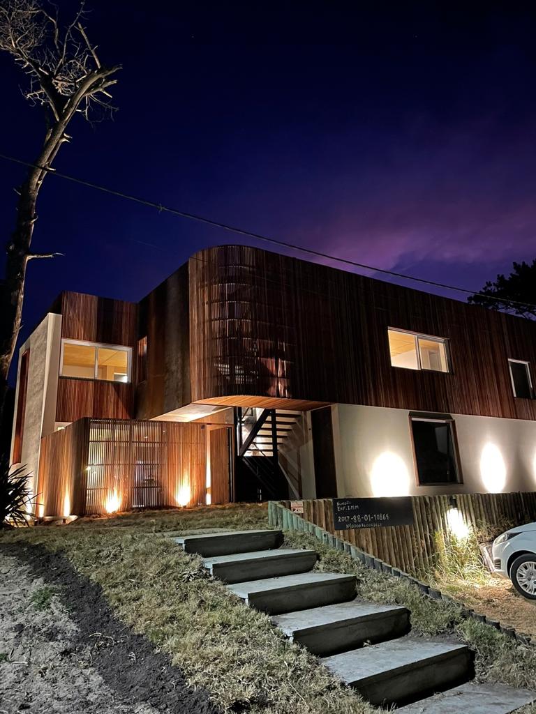 A modern house is illuminated at night, featuring a combination of wood and white facades. Soft outdoor lighting highlights the stairs leading up to the entrance. Large windows allow glimpses of the interior. A tree is visible on the left side, enhancing the natural surroundings.