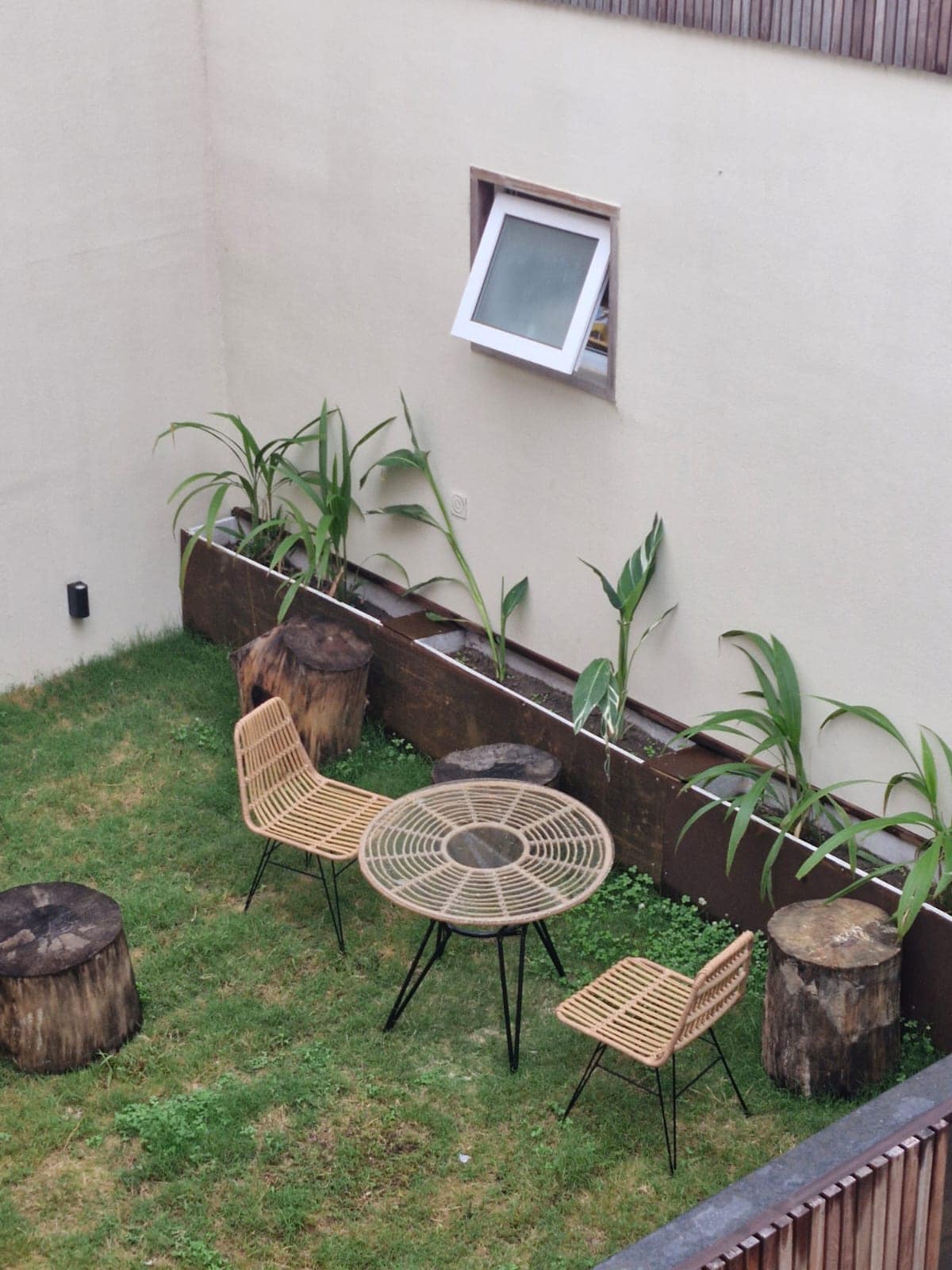 An outdoor garden space features a round table surrounded by four rattan chairs, positioned on a grassy area. Natural wood stumps serve as additional seating or decor. Plants line the edges, contributing to a serene and natural atmosphere.