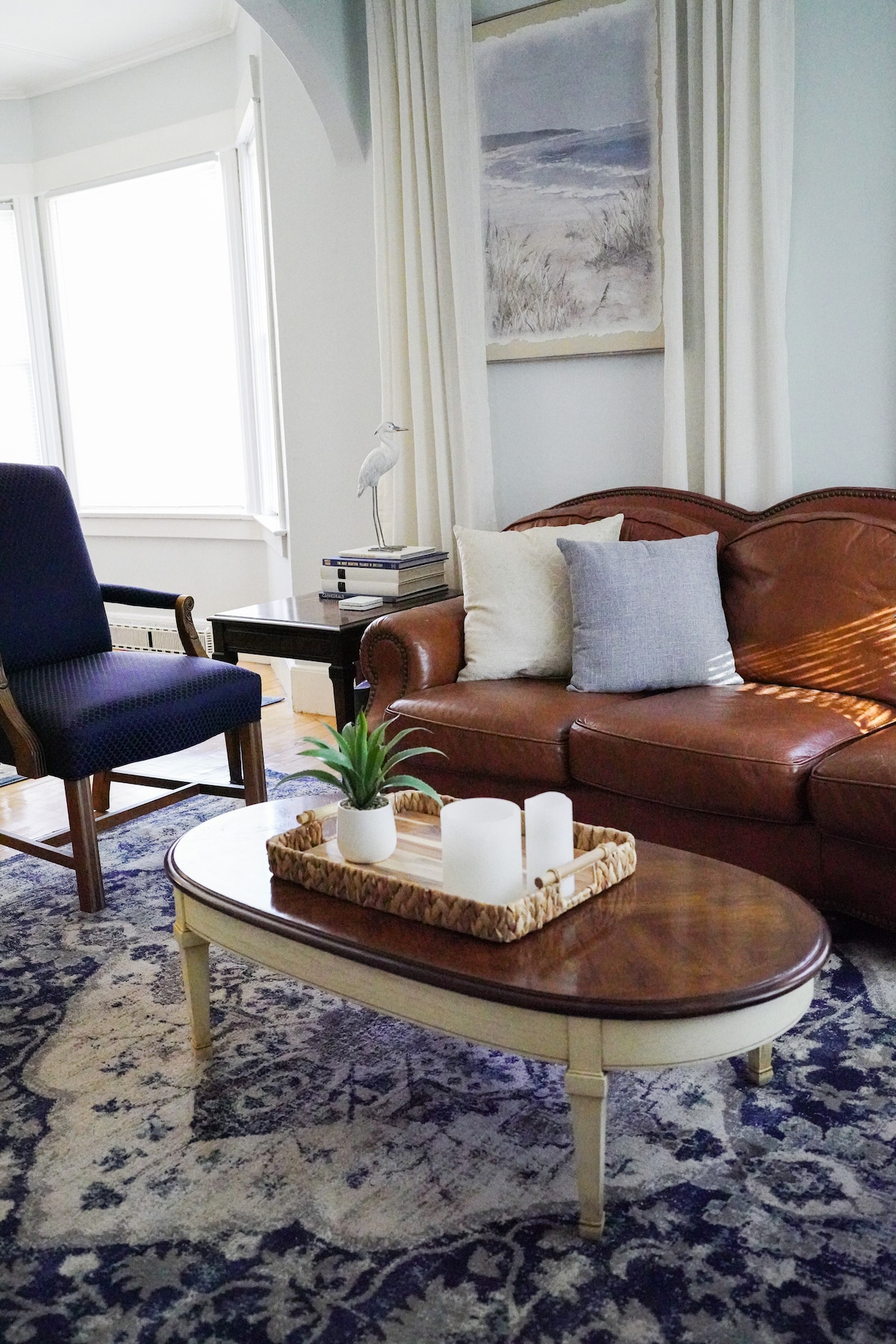 A living room features a brown leather sofa adorned with decorative pillows, situated on a patterned rug. A wooden coffee table with a tray holds three white candles and a small plant. Natural light filters through the surrounding windows, creating a serene atmosphere.