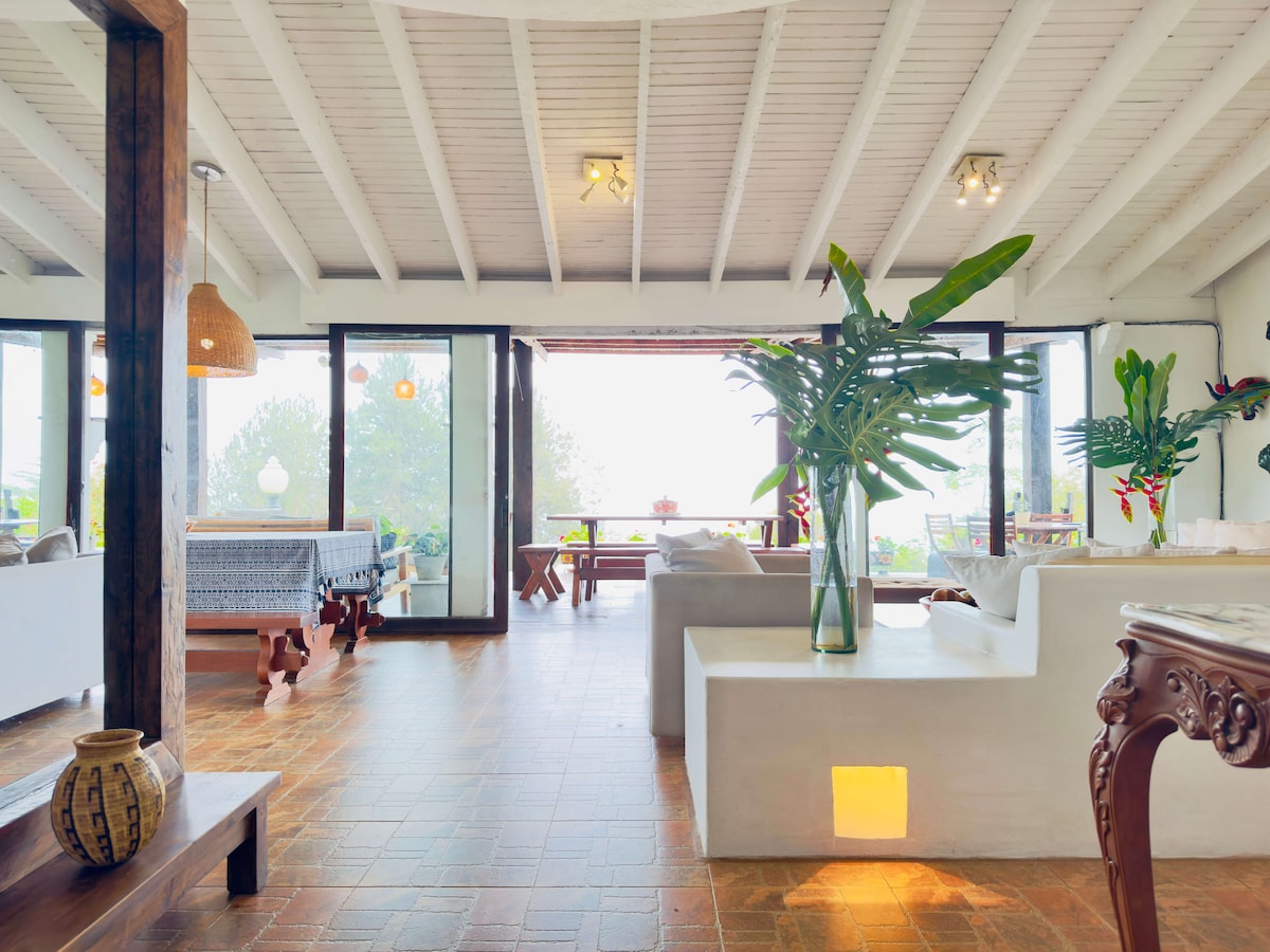 A spacious living area features large glass doors that open to the outdoor view. Natural light fills the room, highlighting the neutral decor and vibrant green plants. A cozy seating arrangement is complemented by a wooden table and rustic accents, providing a welcoming ambiance.