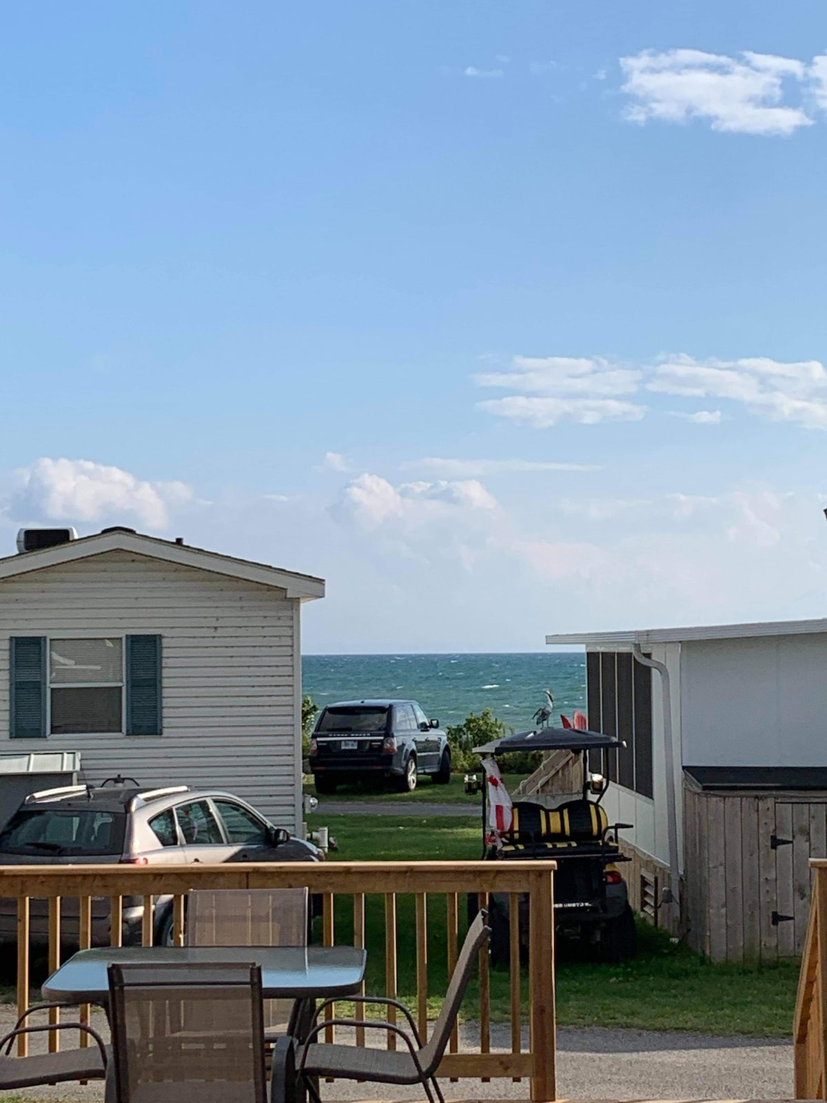 A view of Lake Erie is captured between two adjacent houses, showcasing the water in the distance. A wooden deck is visible in the foreground with patio furniture, while vehicles and a golf cart are parked nearby. The sky features scattered clouds.
