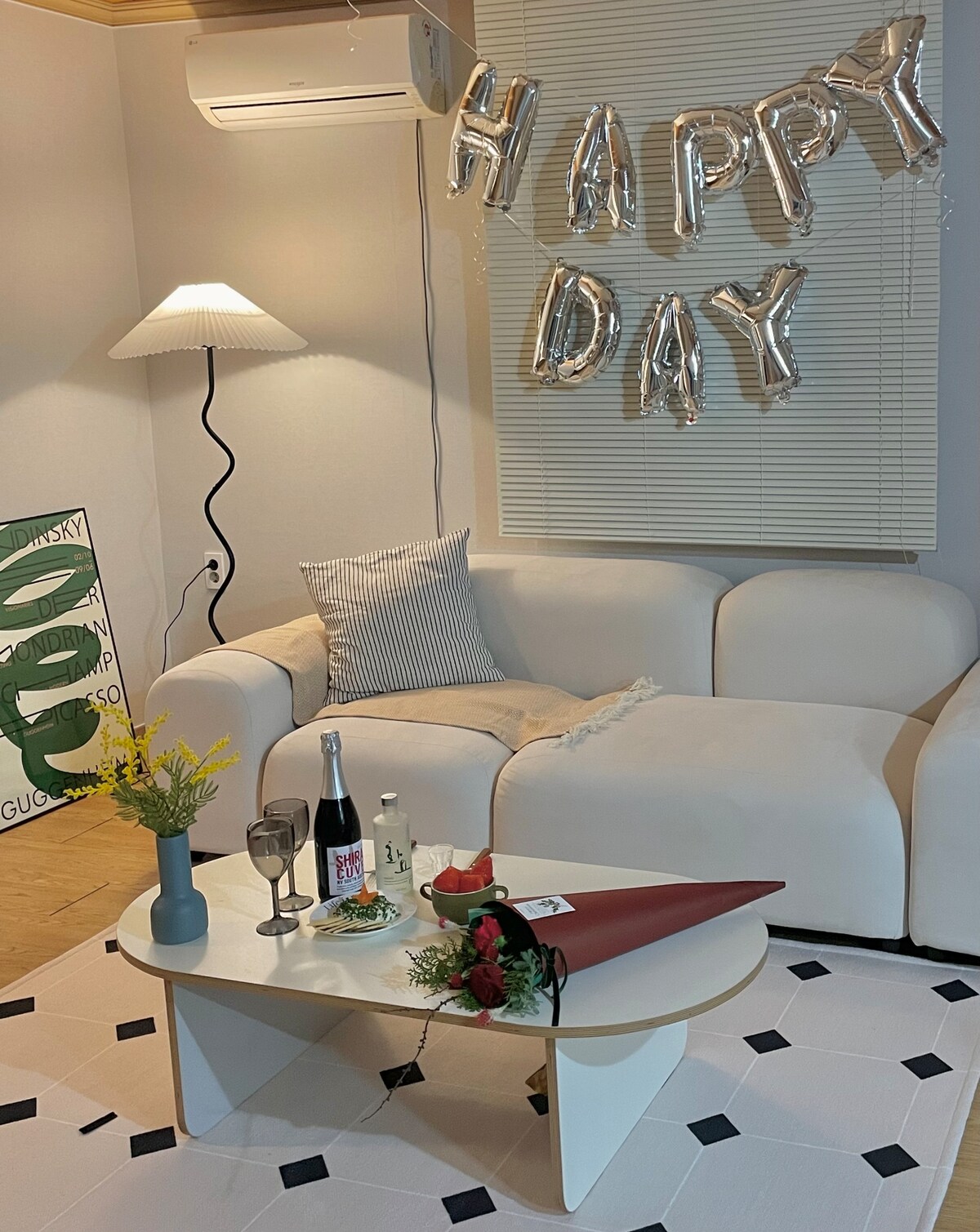 A cozy living area is presented, featuring a light-colored sofa adorned with a decorative throw. A small table is set with two glasses, a bottle of sparkling beverage, and floral arrangements. Metallic balloons spell out 'HAPPY DAY' above, enhancing the celebratory atmosphere.