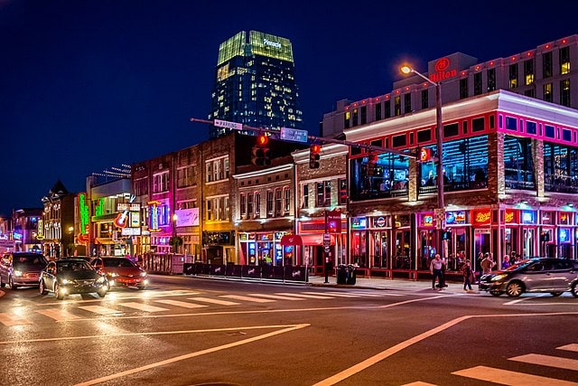 A bustling street is illuminated by vibrant neon lights from various establishments. The scene showcases a mix of vehicles navigating an intersection, while pedestrians are seen on the sidewalks. A tall building in the background is softly lit, adding to the lively atmosphere.