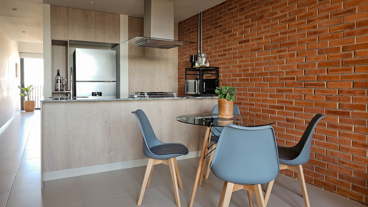 A modern dining area features a round glass table surrounded by four sleek, light blue chairs. A brick accent wall adds texture to the space, while a small potted plant enriches the atmosphere. The open kitchen behind includes stainless steel appliances.