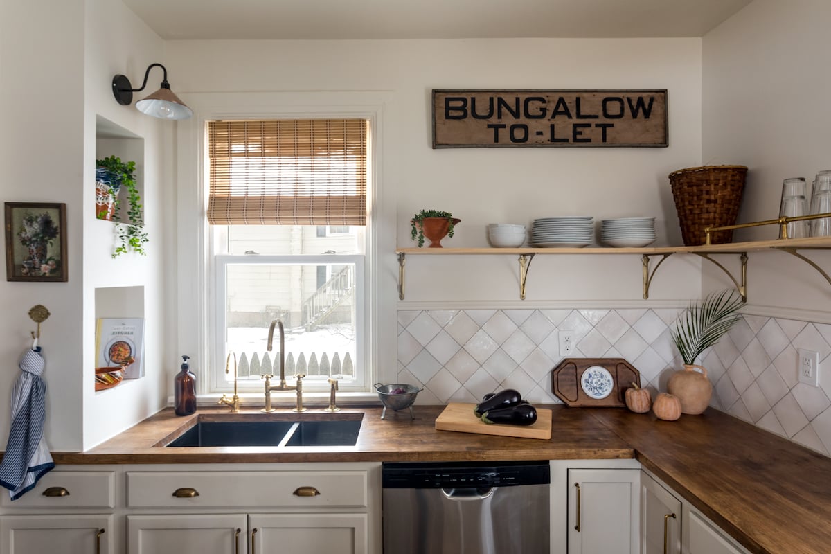 The kitchen space features wooden countertops complemented by warm tones. A double sink is positioned below a window that offers natural light, and shelving displays dishes and decor. A modern dishwasher and a coffee maker are visible, enhancing the functional appeal of the area.